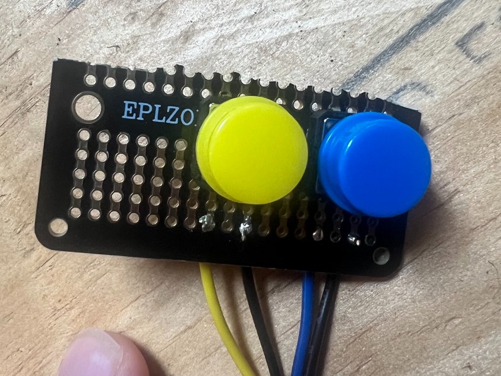 A yellow and a blue push button soldered to a small black prototyping board, with four wires coming off the bottom