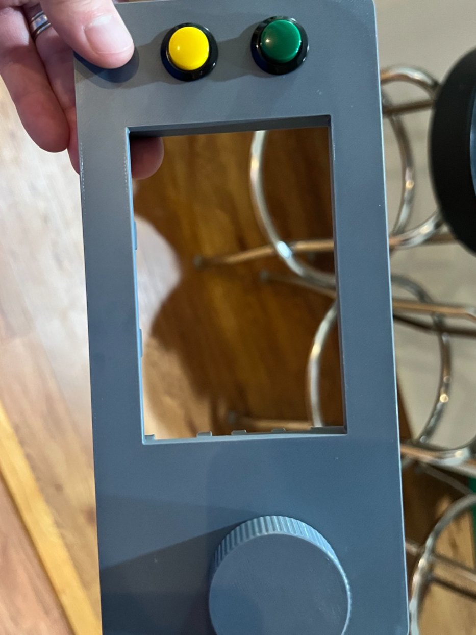 A gray 3D-printed faceplate held in one hand, with a yellow and a green button at one end, an empty screen window, and a knob at the other end