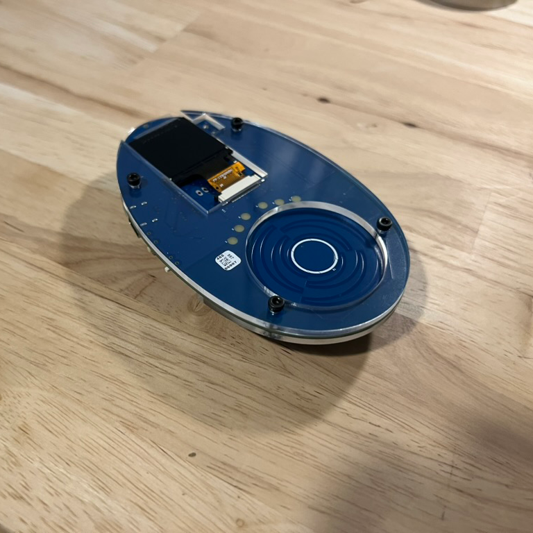 THOTCON 0xD badge (2025): oval blue board with a screen and touch wheel under clear acrylic