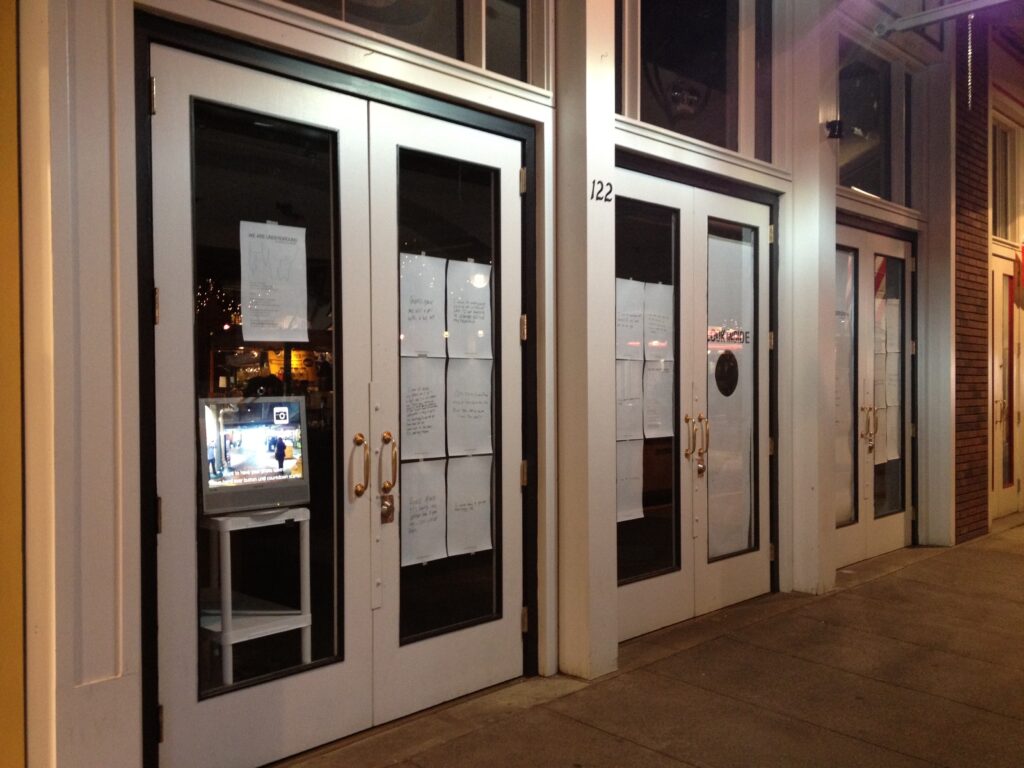 The front of a store with glass windows and doors papered over by notes. A computer monitor sits in the nearest window, waiting to photograph passers-by.